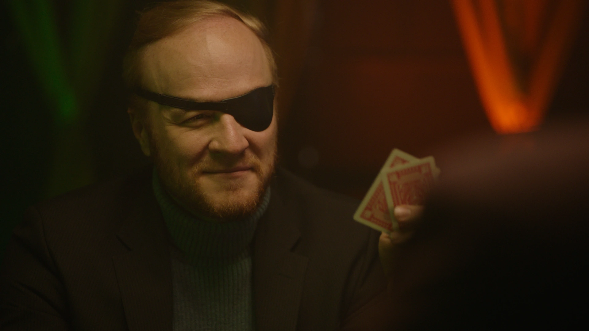 A man wearing an eyepatch holding playing cards in warm, low light